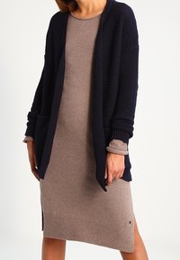 Námořnický pletený cardigan s otevřeným předním dílem, texturovanou tkaninou a žebrovanými rukávy spárovaný s přiléhavým béžovým šaty s bočními rozparky.