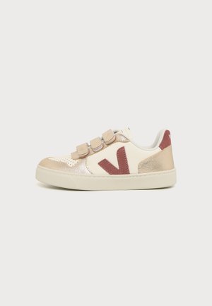 Baskets basses avec panneaux beige, crème et or métallique, trois bandes velcro beige et un logo "V" rouge foncé sur le côté.