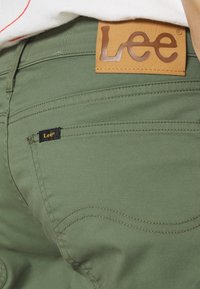 Pantalon en tissu vert olive avec une étiquette en cuir marron à la taille portant "Lee" en lettres embossées. Comprend une petite étiquette noire Lee sur la poche.