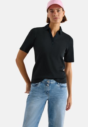 Kvinde iført en pink kasket, sort kortærmet skjorte med krave og lyseblå jeans, stående med den ene hånd hvilende på hoften.