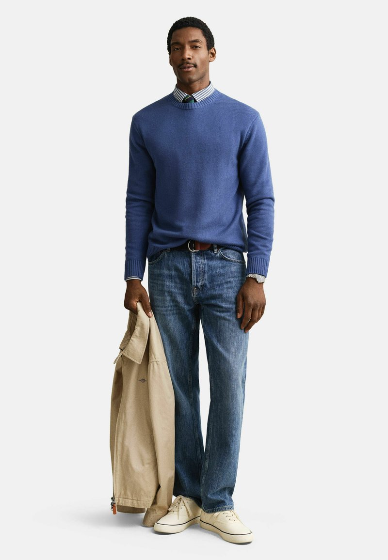 Mann trägt blauen Pullover, gestreiftes Hemd mit Krawatte, Jeans und weiße Turnschuhe, hält in seiner linken Hand eine beige Jacke.