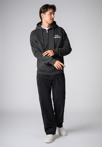 Black zip-up hoodie with drawstrings and front graphic, paired with dark jeans and white sneakers. Smooth fabric and casual fit.