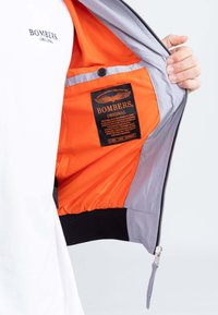 Giacca bomber grigia con fodera arancione brillante e orlo nero. Presenta un'etichetta con testo ed emblema, chiusura a zip e tasca interna.