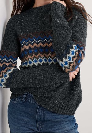 Dunkelgrauer gestrickter Pullover mit blauen, braunen und weißen Zickzackmustern über Brust und Ärmeln, kombiniert mit blauen Jeans.