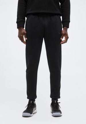 Homme portant un pantalon de jogging noir, un haut à manches longues noir et des chaussures de sport noires, debout sur un fond clair uni.