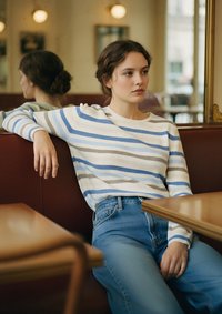 Jersey de rayas en crema, azul y gris; corte relajado. Jeans azules de tiro alto. El entorno incluye una mesa de madera y asientos marrones.