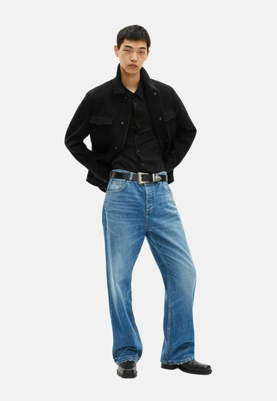 Giacca nera, camicia a fantasia, jeans blu a gamba larga, fibbia argentata, scarpe nere, posato contro uno sfondo bianco semplice.