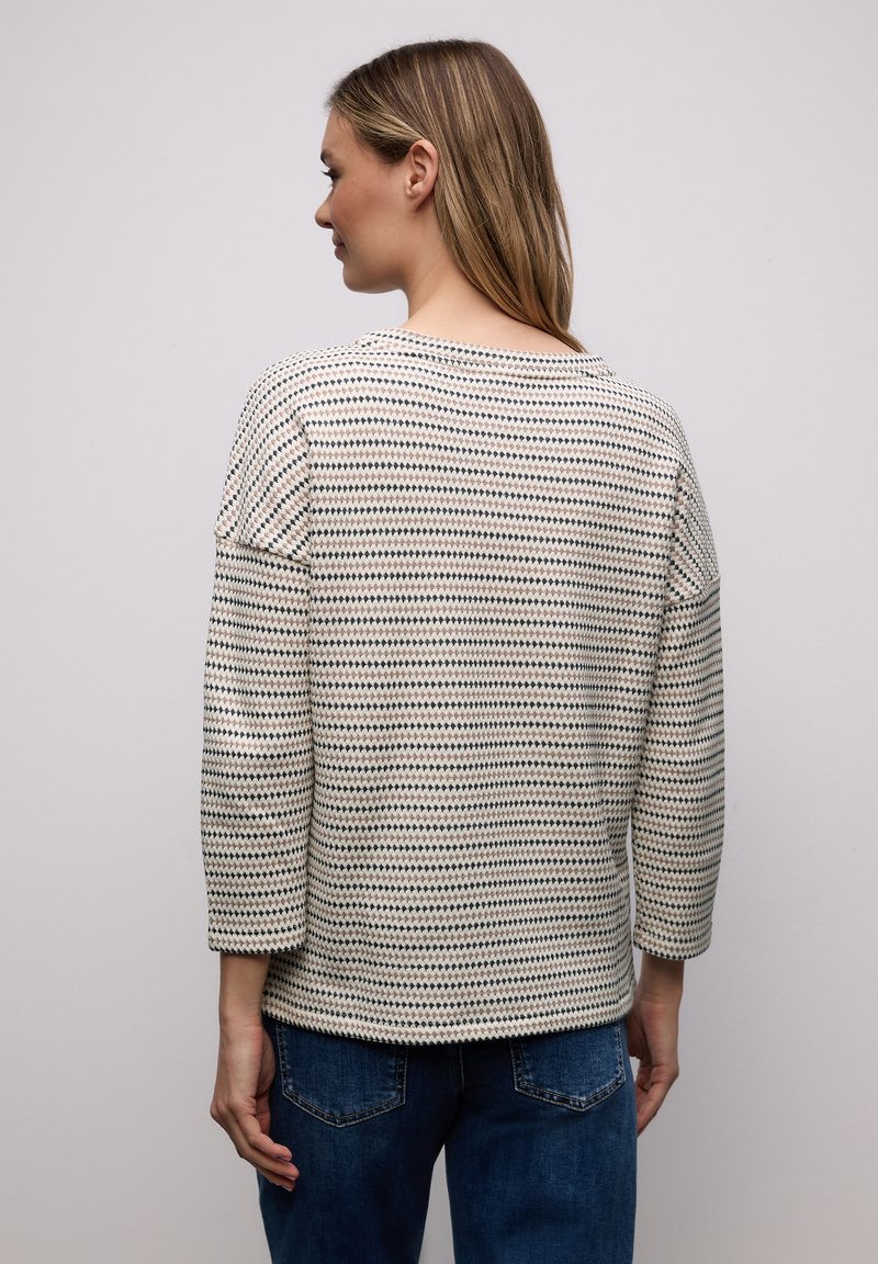 Femme aux cheveux mi-longs portant un haut à manches longues à motifs blancs et noirs et un jean bleu, vue de dos sur un fond uni.