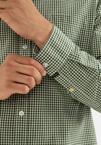 Camisa a cuadros verde y blanca con cuello abotonado, que presenta botones transparentes y una pequeña etiqueta de logo en el puño.