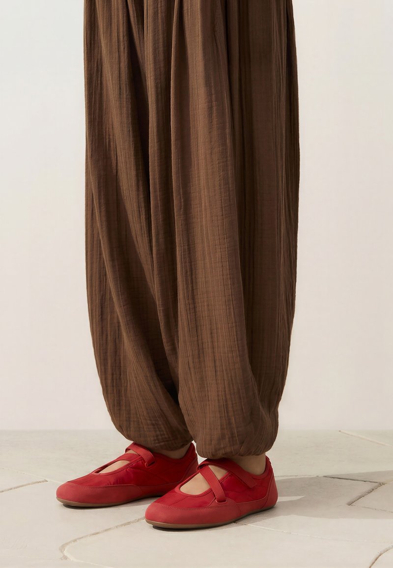 Personne portant un pantalon marron ample et des chaussures à brides rouges debout sur un sol carrelé.