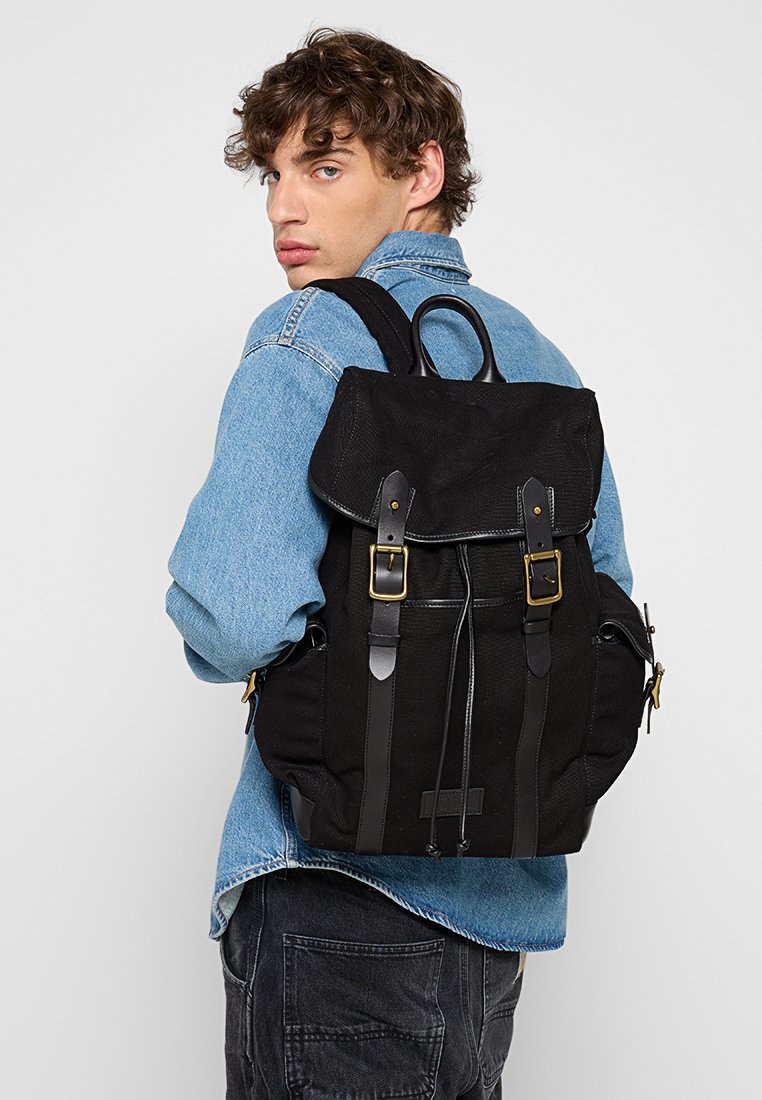 Schwarzer Canvas-Rucksack mit Lederakzenten, verstellbaren Trägern und goldenen Beschlägen. Verfügt über einen Klappenverschluss und eine strukturierte Oberfläche.