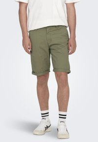 Shorts en coton vert olive avec une coupe regular, ourlets roulés, poches latérales, associés à des baskets blanches et des chaussettes à rayures noires.