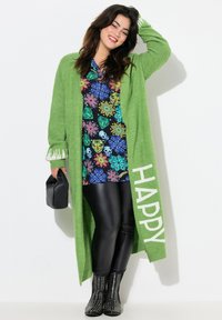 Cárdigan largo verde con el texto en blanco "HAPPY", superpuesto a una camisa negra de flores. Los leggings negros de piel sintética y las botas negras con tachuelas completan el look.