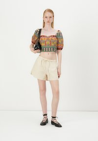 Värviline lühike blousee puffed varrukatega, keerukate mustritega, koos beežide lühikestega ja mustade nööritavate korteritega, hoides käes musta kotti.