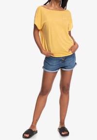 Keltainen lyhythihainen paita, jossa on leveä kaula-aukko, yhdistettynä sinisiin farkkushortseihin, joiden lahkeet on käännetty. Mallilla on päällään mustat liukastettavat kengät, joissa on logotiedot.