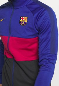 Nike track jacket featuring purple, pink, and black color blocks. Includes embroidered FC Barcelona logo and gold Nike swoosh. High collar.