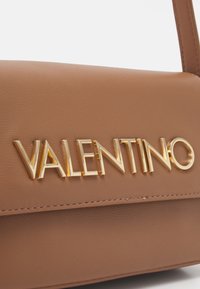Borsa a mano in pelle marrone con una texture liscia e una forma strutturata. Presenta un logo "VALENTINO" dorato in evidenza e cuciture dettagliate.