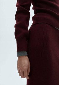 Pull bordeaux à texture côtelée, avec des manches longues et des accents de poignets gris. Gros plan sur une main reposant sur une jupe bordeaux assortie.