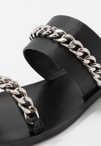Svart sandal med bred texturerad rem och metallkedjeaccent. Slät sula och minimalistisk design som betonar enkelhet och kontrast.