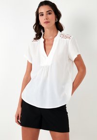 White short-sleeve blouse with a V-neck, featuring lace accents on shoulders and a gathered detail at the chest, paired with black shorts.