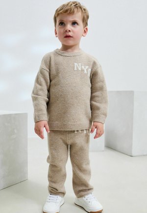 Beige gebreide trui met "NY" logo, bijpassende broek en witte sneakers. Zachte textuur, losse pasvorm, ronde halslijn, ribgebreide mouwen en zoom.