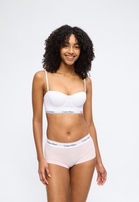 Γυναίκα με σγουρά μαλλιά που χαμογελά, φοράει λευκό σουτιέν Calvin Klein και ανοιχτό ροζ σλιπ Calvin Klein τύπου boyshort, στέκεται μπροστά σε απλό φόντο.