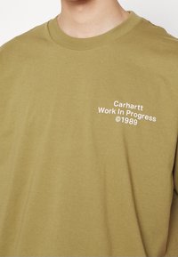 Camiseta de algodón verde oliva con cuello redondo, mangas cortas y texto blanco que dice "Carhartt Work In Progress ©1989" en el pecho.