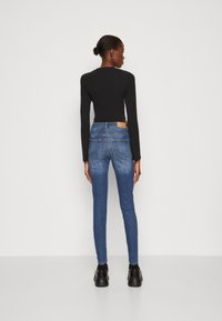 Μαύρη ριχτή μπλούζα με μακριά μανίκια συνδυασμένη με μπλε στενές τζιν, με δύο πίσω τσέπες και ένα καφέ δερμάτινο λογότυπο στη μέση. Μαύρα παπούτσια.