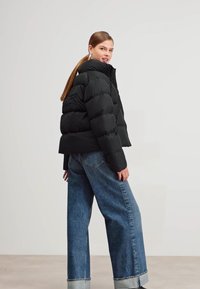 Giubbotto nero in piuma con design corto, texture trapuntata, colletto alto e maniche ampie, abbinato a jeans di denim blu larghi con orli risvoltati.