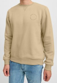 Beige sweatshirt lavet af blødt stof, med rund halsudskæring, ribbede manchetter og et lille rundt logo på brystet.