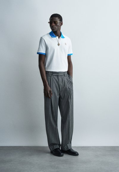 Άνδρας που φοράει λευκό πόλο με μπλε γιακά και μανσέτες, ριγέ φαρδιά παντελόνια, μαύρα παπούτσια, γυαλιά ηλίου και κολιέ με μενταγιόν, στέκεται μπροστά σε γκρι τοίχο.
