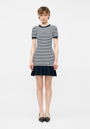 Mujer de pie con un vestido ajustado de rayas horizontales en blanco y negro, mangas cortas y bajo negro sólido con volantes.