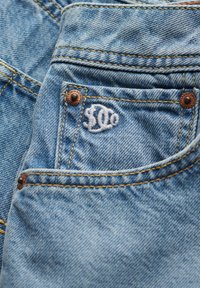 Jeans de mezclilla en azul claro con un pequeño logo bordado en el bolsillo delantero, remaches de cobre y costuras amarillas en contraste.