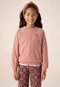 Sweat-shirt en polaire rose avec un col côtelé, arborant un logo en forme de cœur rouge. Associé à un pantalon bordeaux à motif avec des designs complexes.