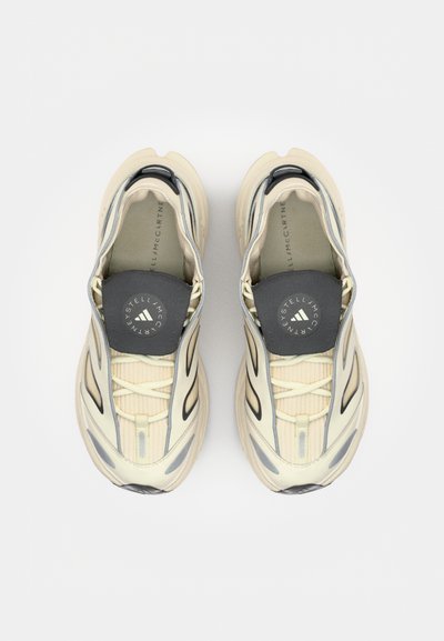 Zapatillas de running en color crema y gris con una textura acanalada y detalles en negro. Cuentan con cordones elásticos y un logo en la lengüeta acolchada.