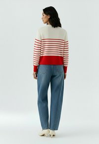 Femme de dos portant un pull rayé blanc et rouge, un jean taille haute bleu et des chaussures blanches à talons sur un fond uni.