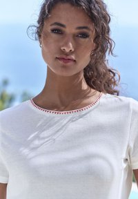 Balta īsām piedurknēm t-kreklā ar apaļu kakla izgriezumu, kuram ir sarkani izšūti akcenti. Mīksta, gluda tekstūra un brīva piegriezuma.