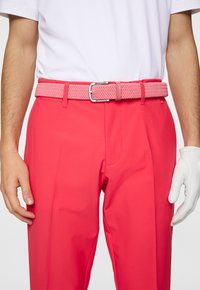 Roze golfbroek met een gladde textuur, voorzien van een gevlochten roze riem en een wit shirt met korte mouwen. Handschoenen aan de linkerhand.