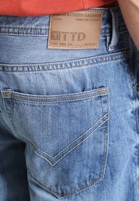 Jeans i denim med en ljusblå tvätt, med en läderlogotyp med texten "TTD" och kontrasterande sömnad på bakfickan.