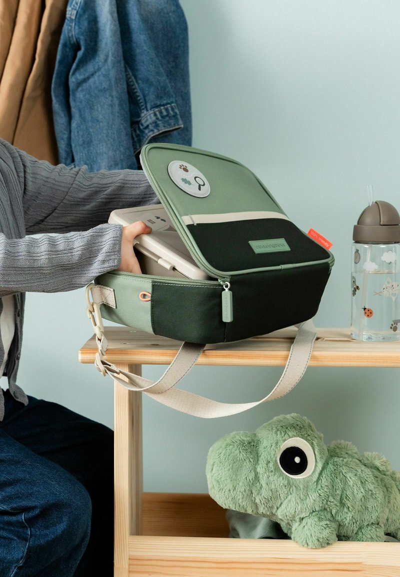 Les mains d’un enfant déposent une boîte à lunch dans un sac vert et noir sur une table en bois, à côté d’un gobelet d’apprentissage et d’un dinosaure en peluche vert.