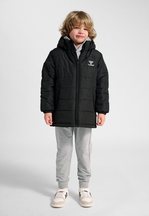 Barn med krøllet blondt hår iført en sort hættetrøje i dunjakke, grå joggingbukser og hvide sneakers stående mod en ensfarvet baggrund.