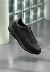 Chaussures de sport noires présentant un mélange de surfaces lisses et texturées, un design à lacets, et une semelle en caoutchouc avec des éléments antidérapants.