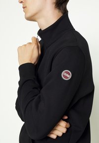 Svart zip-up fleecejacka med hög krage, ribbade manschetter och en rund logotypapplikation på ärmen. Slät textur och figurnära design.