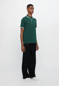 Ung mann står oppreist, iført en mørkegrønn polo-skjorte med hvit kant, sorte vide bukser og hvite sneakers foran en ensfarget hvit bakgrunn.