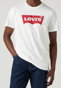 Man wearing white short-sleeve Levi's t-shirt with red logo, paired with dark blue denim jeans, hand in pocket.