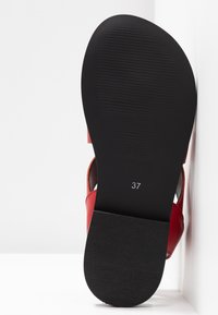 Punainen nahkasandaali, jossa on teksturoitu musta kumipohja. Koko 37 merkitty pohjaan. Ominaisuuksiin kuuluu minimalistinen muotoilu avointen hihnojen kanssa.