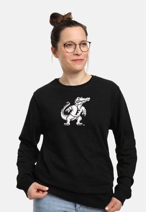 Kobieta w okularach i włosach upiętych w kok, ubrana w czarną bluzę z białym karykaturalnym aligatorem trzymającym piłkę do futbolu amerykańskiego z przodu.