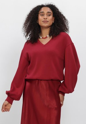 Woman with curly hair wearing a loose red V-neck sweater and matching skirt, standing against a plain background.