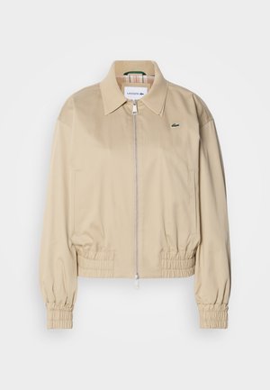 Chaqueta corta beige hecha de tejido ligero, con cremallera completa, puños elásticos y un logo discreto en el pecho.