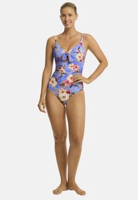 Glunderende vrouw in een badpak met bloemenprint staat met één hand op haar heup en toont de helderblauwe stof tegen een witte achtergrond.